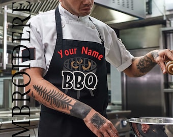 May include: Black apron featuring the word "BBQ" in white block letters and a grill graphic. The apron has the text "Your Name" in red script above the grill graphic. The word "EMBROIDERED" is printed vertically on the left side.