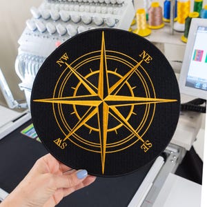 May include: A circular black embroidered patch featuring a gold compass rose design. The compass includes cardinal direction markers for NW, NE, SE, and SW in gold lettering. The design consists of a central star with radiating points.