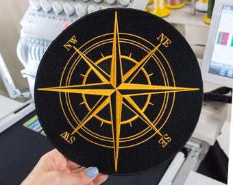 May include: A circular black embroidered patch featuring a gold compass rose design. The compass includes cardinal direction markers for NW, NE, SE, and SW in gold lettering. The design consists of a central star with radiating points.