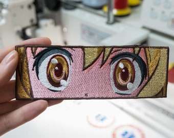 May include: Embroidered patch featuring a close-up of a pair of anime eyes with brown irises and white pupils. The eyes are surrounded by blonde hair and a pink background.
