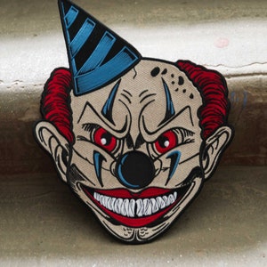 May include: A creepy embroidered patch of a clown with a red and blue hat, red hair, a black nose, and a wide, toothy grin.