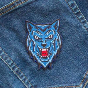 Puede incluir: Parche bordado de cabeza de lobo azul con una expresión feroz y dientes afilados. El parche está sobre un fondo de mezclilla.