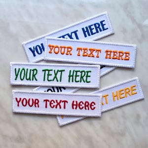 Może przedstawiać: Prostokątne, białe, haftowane naszywki z napisem "YOUR TEXT HERE" w różnych kolorach: niebieskim, pomarańczowym, zielonym, czerwonym i żółtym. Każda naszywka ma białą obwódkę i jest prezentowana na marmurowej powierzchni.