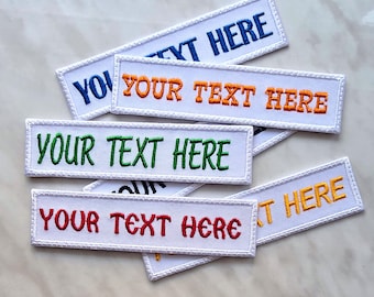 May include: A collection of rectangular white embroidered patches. Each patch features the text "YOUR TEXT HERE" in different colours, including blue, orange, green, red, and yellow, with a white border. Displayed on a marble surface.