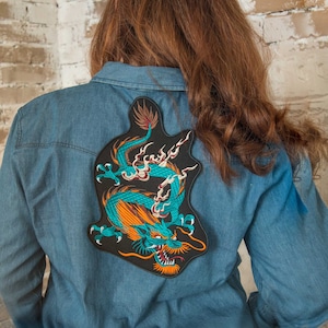 Könnte beinhalten: Ein blaues Jeanshemd mit einem gestickten Aufnäher eines grünen Drachen mit orangefarbenen Akzenten auf der Rückseite.