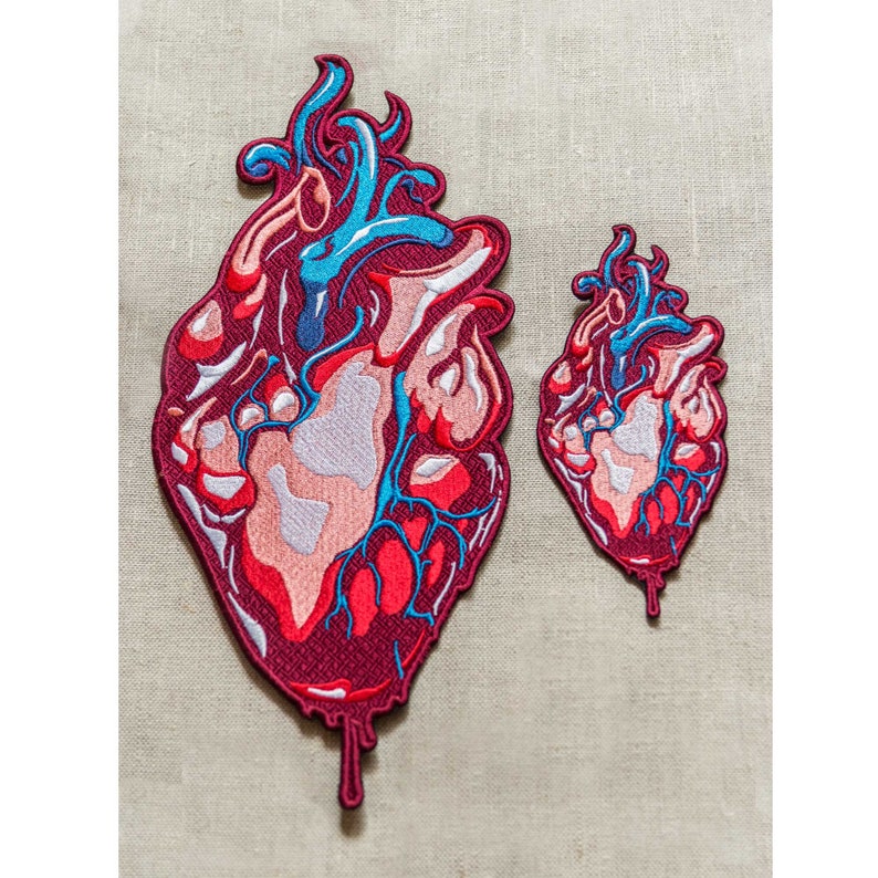 Op de afbeelding: Twee geborduurde patches van een rood, roze en blauw anatomisch hart met druppelend bloed. De grotere patch is ongeveer 12,5 cm hoog en de kleinere ongeveer 7,5 cm hoog.