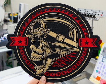 May include: A circular embroidered patch featuring a gold skull wearing a helmet with goggles. The design is framed by a red flame border and a red chain link circle, with two red banners on the sides each displaying a single black star.