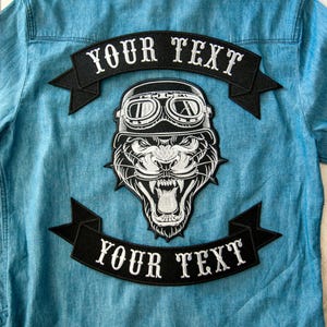 May include: A light blue denim shirt with a large black and white patch featuring a roaring tiger wearing a helmet and goggles. The patch has the text "YOUR TEXT" on two black banners.