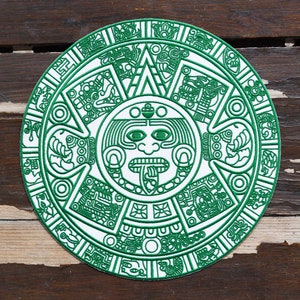 May include: A circular embroidered patch featuring a white background with intricate green thread detailing. The design depicts a central face with a protruding tongue surrounded by concentric rings of geometric patterns and stylized figures.