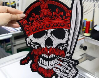 May include: An embroidered patch featuring a skull wearing a red crown with fleur-de-lis details. The skull has a thick red beard and is holding a long, silver-white sword. The design uses black, red, and white thread with a black border.