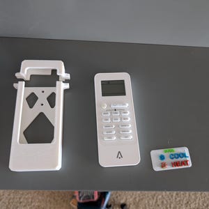 May include: White air conditioner remote control, bracket, and a small rectangular sticker with the words "COOL" and "HEAT" in blue, green, and red. The remote has multiple buttons and a small display screen. The bracket has a unique geometric design.