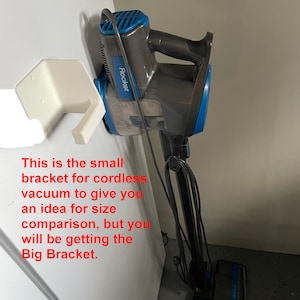 Puede incluir: Una aspiradora inalámbrica azul y gris está montada en un soporte de pared blanco. La aspiradora está etiquetada como "Rocket". El texto "This is the small bracket for cordless vacuum to give you an idea for size comparison, but you will be getting the Big Bracket." está escrito debajo de la imagen.