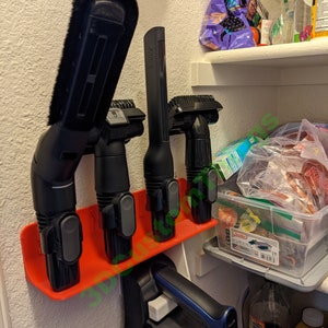 May include: A red plastic wall-mounted organizer with four black vacuum cleaner attachments. The attachments include a crevice tool, a dusting brush, an upholstery tool, and a combination tool. The organizer is mounted on a white wall.