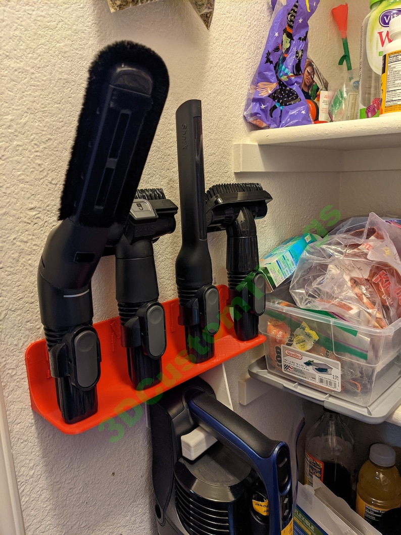 May include: A red plastic wall-mounted organizer with four black vacuum cleaner attachments. The attachments include a crevice tool, a dusting brush, an upholstery tool, and a combination tool. The organizer is mounted on a white wall.