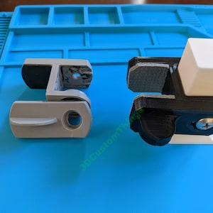 May include: Two 3D printed plastic clamps, one gray and one black, with a white plastic base. The black clamp has a screw on the side.