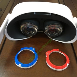 Puede incluir: Un auricular de realidad virtual blanco con dos lentes negros. Se muestran dos placas faciales de repuesto debajo del auricular, una azul y una roja.