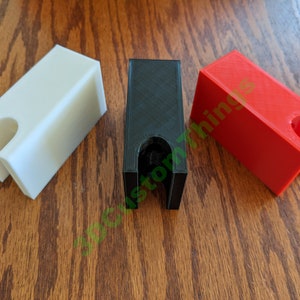 Puede incluir: Tres soportes de plástico impresos en 3D en blanco, negro y rojo. Cada soporte tiene una forma rectangular con una abertura circular en el centro.