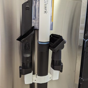 Puede incluir: Soporte de pared blanco con tres accesorios de aspiradora negros. Los accesorios están etiquetados como "CORDLESS" y "Shark".