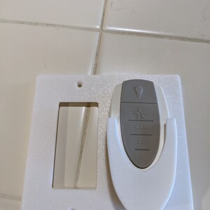 May include: White plastic wall plate with a cutout for a light switch and a holder for a remote control. The remote control is gray and has a light bulb icon, a star icon, and several buttons.