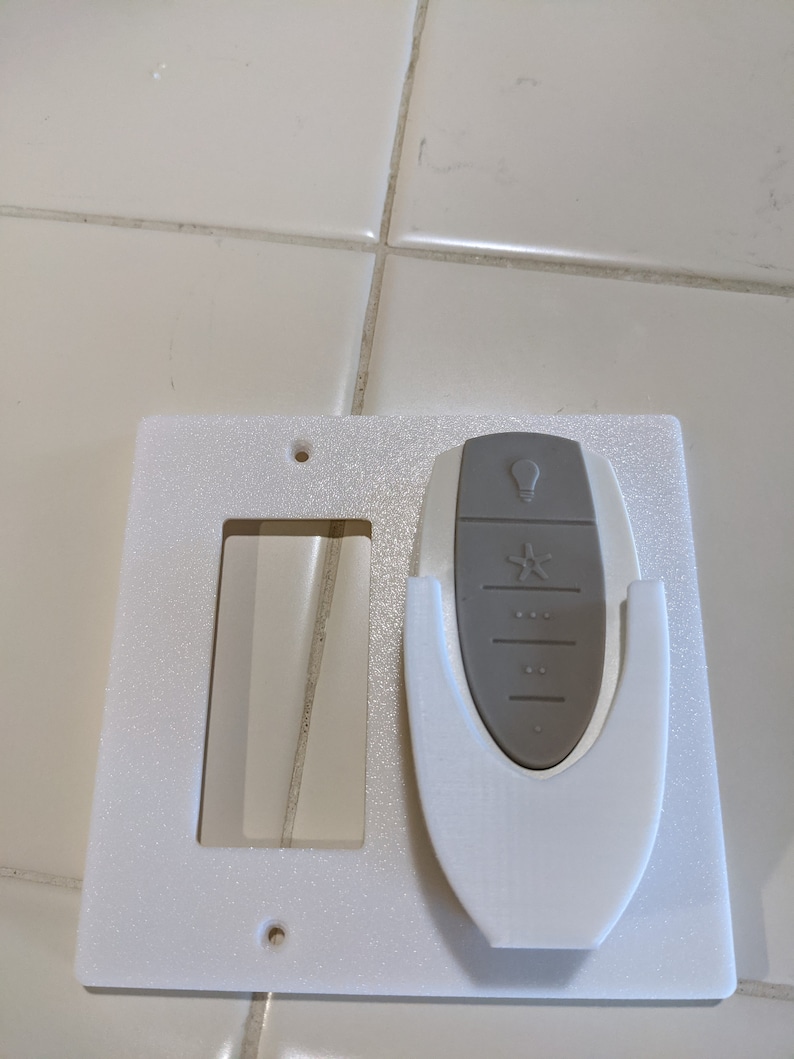 May include: White plastic wall plate with a cutout for a light switch and a holder for a remote control. The remote control is gray and has a light bulb icon, a star icon, and several buttons.