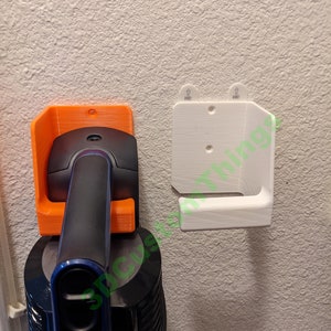May include: Two 3D printed wall mounts for a cordless vacuum cleaner. The mount on the left is orange and the mount on the right is white. Both mounts have a hook for hanging the vacuum cleaner.