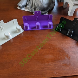 Puede incluir: Tres ganchos de pared impresos en 3D en blanco, morado y negro. Cada gancho tiene tres puntas.