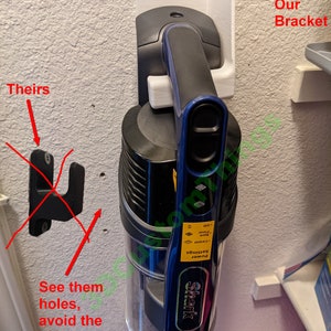 May include: A white 3D printed wall mount bracket for a Shark vacuum cleaner. The bracket is mounted on a white wall. The vacuum cleaner is hanging on the bracket. The vacuum cleaner is blue and black with a silver canister. The vacuum cleaner has a power setting dial with the text "Settings", "Power", "Carpet", "Bare Floor", and "Off". The vacuum cleaner also has the text "Shark" on the side. The image shows a close-up of the vacuum cleaner and the bracket. The image also shows a red arrow pointing to the bracket and the text "Our Bracket". The image also shows a red arrow pointing to a black and white wall mount hanger and the text "Theirs". The image also shows a red arrow pointing to the holes in the wall and the text "See them holes, avoid the mistake, don't use hangers".
