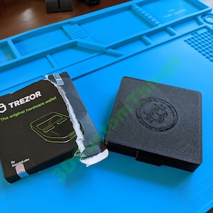 May include: A black Trezor hardware wallet in its original packaging. The box is black with white text that reads "Trezor The original hardware wallet" and a green logo of a lock with a keyhole. The wallet is black with a raised logo of a lock with a keyhole.