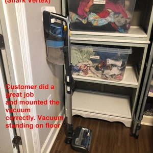 May include: A gray and blue Shark Vertex cordless vacuum cleaner is mounted on a white wall mount. The vacuum is standing on a wood floor.