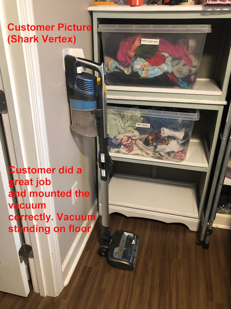 May include: A gray and blue Shark Vertex cordless vacuum cleaner is mounted on a white wall mount. The vacuum is standing on a wood floor.
