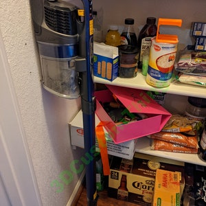 May include: A blue and gray cordless stick vacuum cleaner with a long extension wand is stored in a kitchen pantry. The vacuum is mounted on a wall hook. The pantry shelves are stocked with various food items, including cereal, rice, and beer.