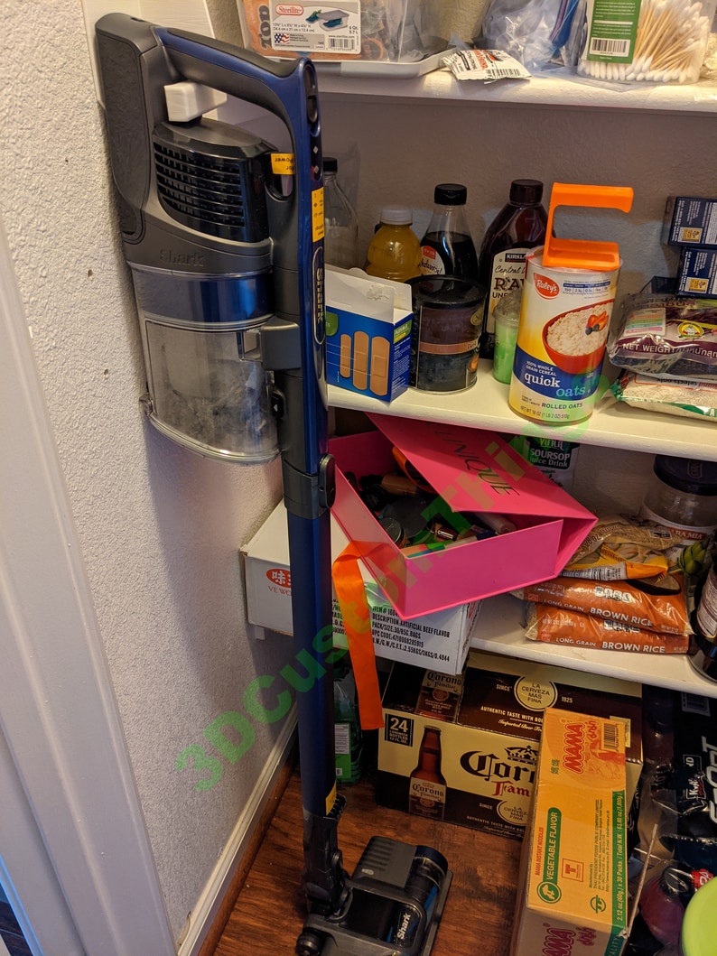 May include: A blue and gray cordless stick vacuum cleaner with a long extension wand is stored in a kitchen pantry. The vacuum is mounted on a wall hook. The pantry shelves are stocked with various food items, including cereal, rice, and beer.