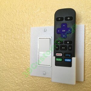 May include: A white plastic remote control holder mounted to a light switch plate. The holder has a slot for a remote control and is designed to hold the remote securely in place. The remote control is black and has buttons for Netflix, Hulu, Sling, and Vudu.
