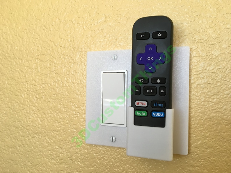 May include: A white plastic remote control holder mounted to a light switch plate. The holder has a slot for a remote control and is designed to hold the remote securely in place. The remote control is black and has buttons for Netflix, Hulu, Sling, and Vudu.