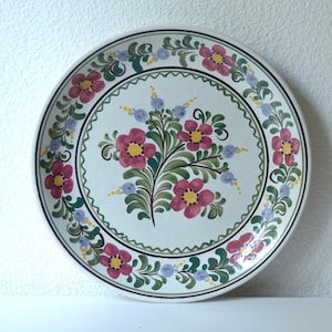 May include: A white ceramic plate with a floral design. The plate features a central bouquet of pink flowers with yellow centers and green leaves. The border includes pink, blue, and green floral accents, and a black and green trim.