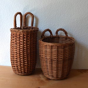 May include: Two brown wicker baskets. One is tall and cylindrical with two arched handles. The other is shorter and round with two small handles. Both are made of woven brown wicker and sit on a wooden surface.