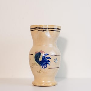 May include: A beige ceramic vase with a rooster design. The vase has a wide base, a narrow neck, and a flared rim. The rooster is depicted in blue and pink, with black outlines. The vase is decorated with horizontal black lines and blue floral accents.