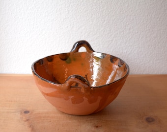 May include: A handmade, terracotta-colored bowl with two arched handles. The bowl has a glossy finish and a dark rim. The interior shows hints of green and black. This rustic bowl is suitable for serving or display.