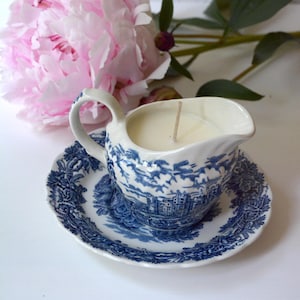Puede incluir: Una pequeña jarra de cerámica azul y blanca llena de una vela, colocada sobre un platillo a juego. La jarra y el platillo presentan un diseño de paisaje detallado. Una gran flor de peonía rosa está en el fondo.