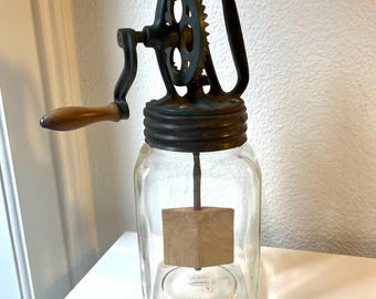 Könnte beinhalten: Eine Vintage-Buttermaschine aus Glas mit einem dunkelgrünen Metallmechanismus und einem Holzgriff. Die Maschine hat ein klares Glasgefäß mit einem Holzpaddel im Inneren. Die Maschine steht auf einer weißen Oberfläche.