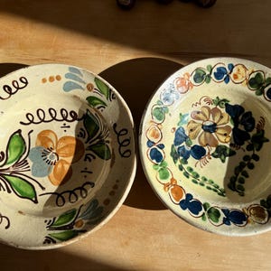 May include: Two round ceramic plates with a cream-colored base and hand-painted floral motifs. The left plate features a central orange and blue flower with green leaves and black scrollwork. The right plate displays a central flower surrounded by a circular border of blue, green, and orange petal-like shapes.