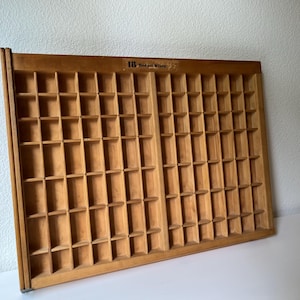 May include: A vintage wooden printer's tray with numerous small compartments. The tray is made of light-colored wood and features the text "HB Badger Mark" at the top. It is designed to hold individual letters or small items.