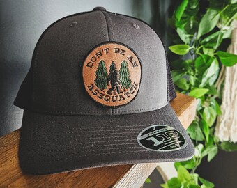 May include: A gray and black trucker hat with a brown leather patch that says "Don't Be An Assquatch" with a green silhouette of a Bigfoot walking through trees.