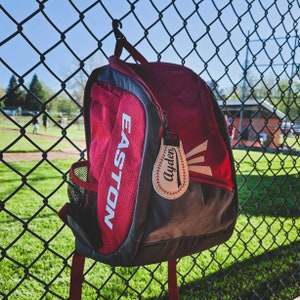 May include: Red and grey baseball backpack with the word "EASTON" printed on the side. A white baseball-shaped keychain with the name "Ayden" is attached to the zipper.