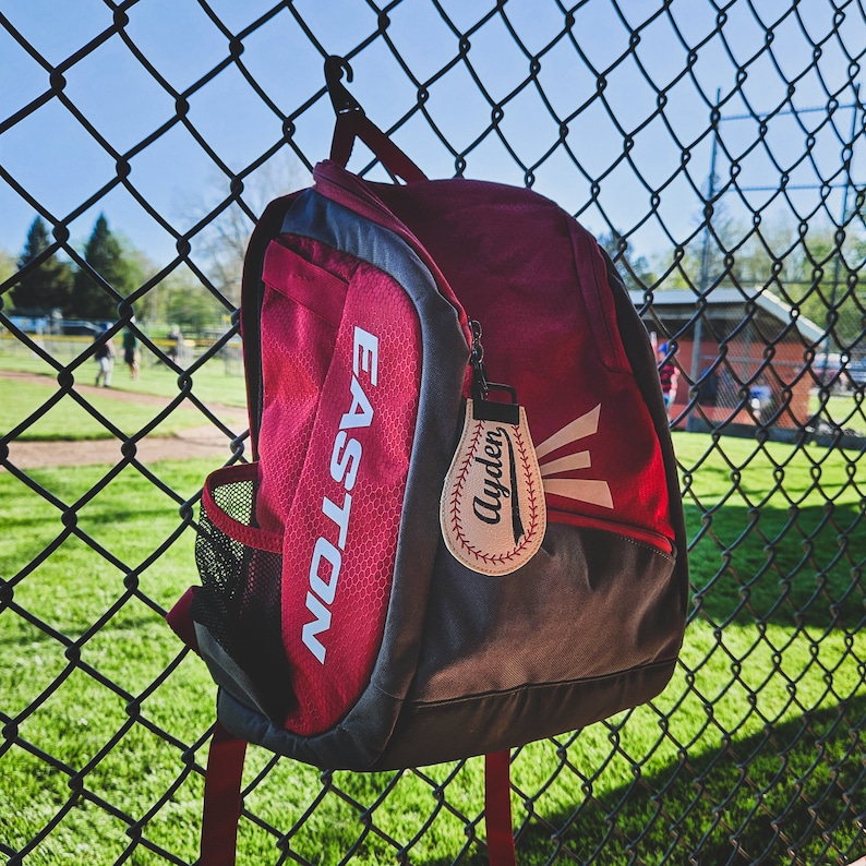 May include: Red and grey baseball backpack with the word "EASTON" printed on the side. A white baseball-shaped keychain with the name "Ayden" is attached to the zipper.
