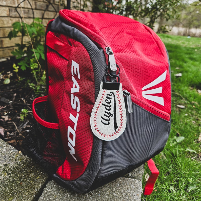 May include: Red and grey baseball backpack with a white baseball-shaped keyring that says "Ayden" on it.