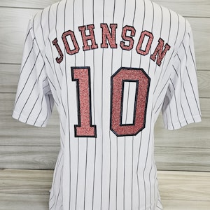 Op de afbeelding: Wit baseballshirt met zwarte strepen. Op de achterkant van het shirt staat de naam "JOHNSON" en het nummer "10" in rood glitter.