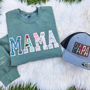 Op de afbeelding: Een groen sweatshirt met het woord "MAMA" gespeld in kleurrijke stofletters. Een blauwe en zwarte baseballpet met het woord "PAPA" gespeld in bruine stofletters.