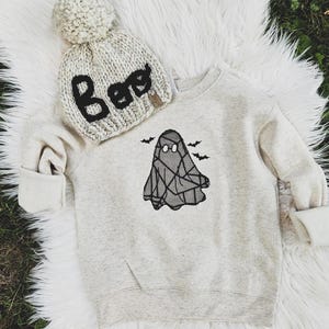 May include: A light gray Halloween-themed sweatshirt with a ghost graphic and three bats. A matching knit beanie with the word "Boo" in black lettering and a pom-pom is also included. The items are displayed on a white fluffy surface.