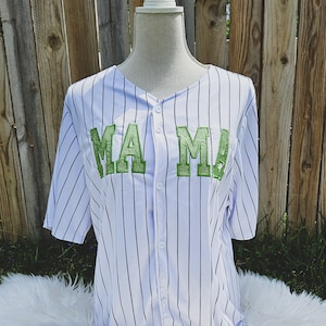 May include: White and black striped baseball jersey with green embroidered lettering that says "MAMA".